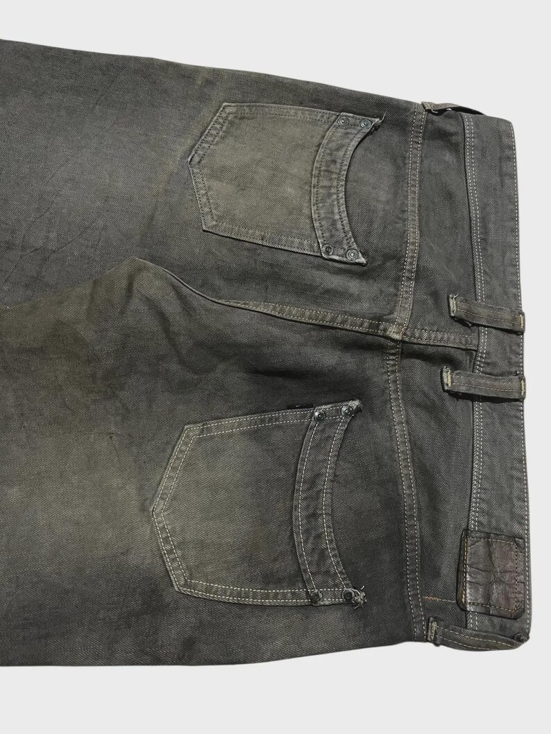 K&ouml;nnte beinhalten: Nahaufnahme der R&uuml;ckseite einer dunkelgrauen Jeans. Die Jeans hat zwei Ges&auml;&szlig;taschen mit dekorativen N&auml;hten und Nieten. Ein kleines Lederpatch ist in der N&auml;he des Bunds sichtbar. Die Jeans ist leicht verblichen, was ihr ein abgenutztes Aussehen verleiht.