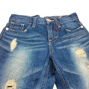 May include: Close-up of blue denim jeans with distressed details. The jeans have a faded wash, visible rips, and white stitching. The front pockets and button closure are visible.