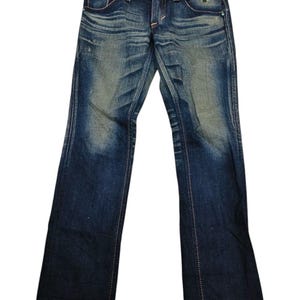 May include: Blue denim jeans with a faded wash and subtle distressing. The jeans feature a classic five-pocket design, a button closure, and a zip fly. White stitching accents the seams and pockets. The jeans have a straight leg fit.