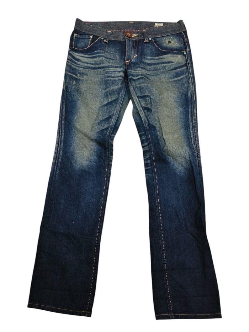 May include: Blue denim jeans with a faded wash and subtle distressing. The jeans feature a classic five-pocket design, a button closure, and a zip fly. White stitching accents the seams and pockets. The jeans have a straight leg fit.