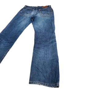 Puede incluir: Vaqueros azules de mezclilla con un dise&ntilde;o cl&aacute;sico de cinco bolsillos. Los vaqueros tienen un lavado descolorido y un parche de cuero en la cintura trasera. Los bolsillos traseros presentan costuras decorativas. Los vaqueros est&aacute;n planos sobre fondo blanco.
