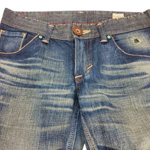 May include: Close-up of blue denim jeans with a faded wash. The jeans have a contrasting gray waistband, decorative stitching, and turquoise accents. A brown button and a small patch detail the front.