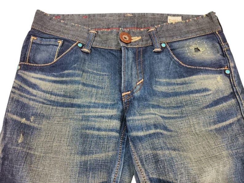 May include: Close-up of blue denim jeans with a faded wash. The jeans have a contrasting gray waistband, decorative stitching, and turquoise accents. A brown button and a small patch detail the front.