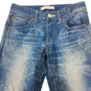 May include: Blue denim jeans with a faded paisley pattern. The jeans have a classic five-pocket design, a button and zipper closure, and belt loops. The fabric has a worn, distressed look.