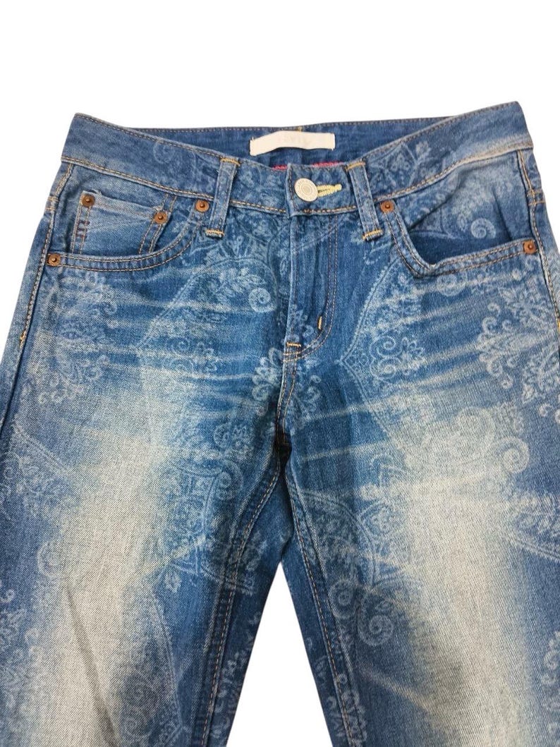 May include: Blue denim jeans with a faded paisley pattern. The jeans have a classic five-pocket design, a button and zipper closure, and belt loops. The fabric has a worn, distressed look.