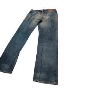 Puede incluir: Vaqueros de mezclilla azul descolorido con un dise&ntilde;o cl&aacute;sico de cinco bolsillos. Los vaqueros tienen un corte de pierna recta y una apariencia desgastada, con decoloraci&oacute;n y desgaste visibles. Un parche de cuero marr&oacute;n es visible en la parte trasera.
