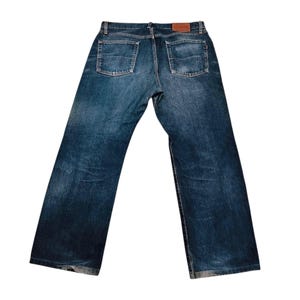 May include: Back view of blue denim jeans with two back pockets and a brown leather patch. The jeans have a classic five-pocket design and a straight leg fit. The denim has a faded wash and a worn appearance.