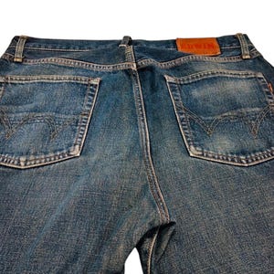 May include: Close-up of the back of blue denim jeans. The jeans have two back pockets with decorative stitching and a brown leather patch with the brand name "EDWIN". The denim has a faded, worn appearance.
