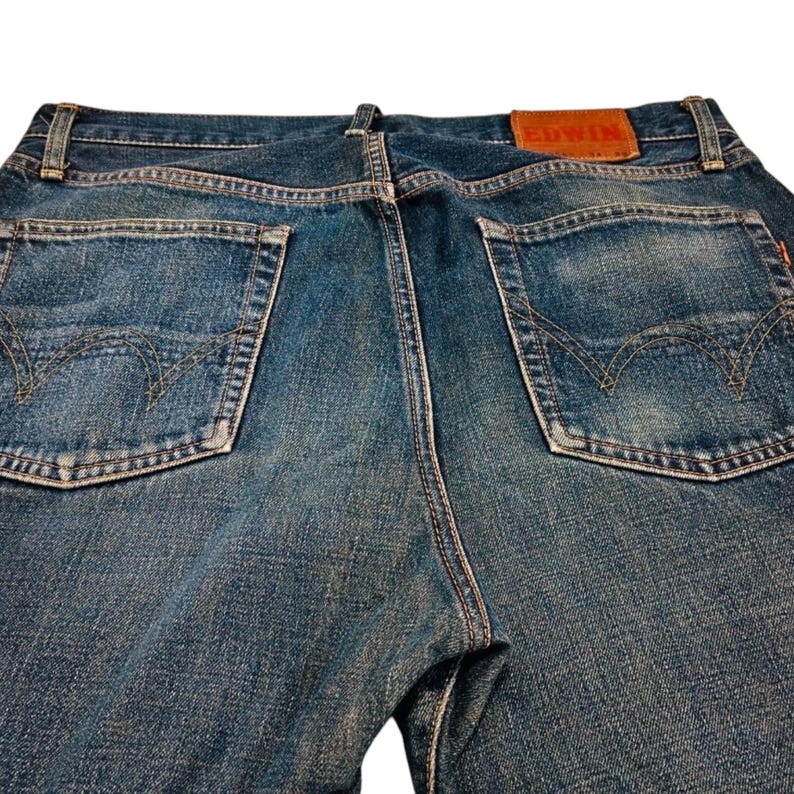 May include: Close-up of the back of blue denim jeans. The jeans have two back pockets with decorative stitching and a brown leather patch with the brand name "EDWIN". The denim has a faded, worn appearance.