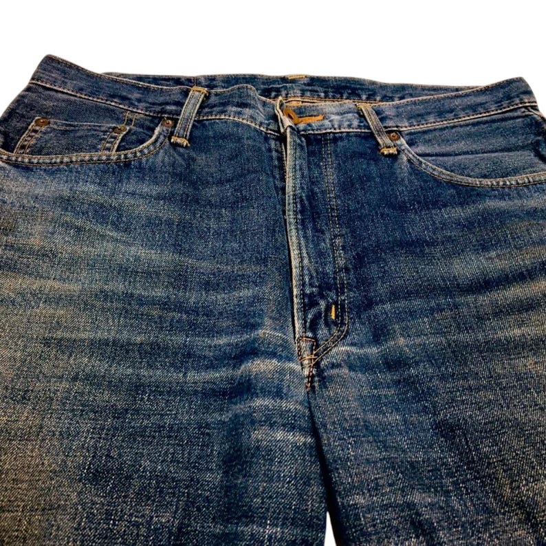 May include: Close-up of blue denim jeans. The jeans have a classic five-pocket design with a button and zipper closure. The denim has a faded, worn appearance, with visible wear marks and creases. The stitching is a contrasting gold color.