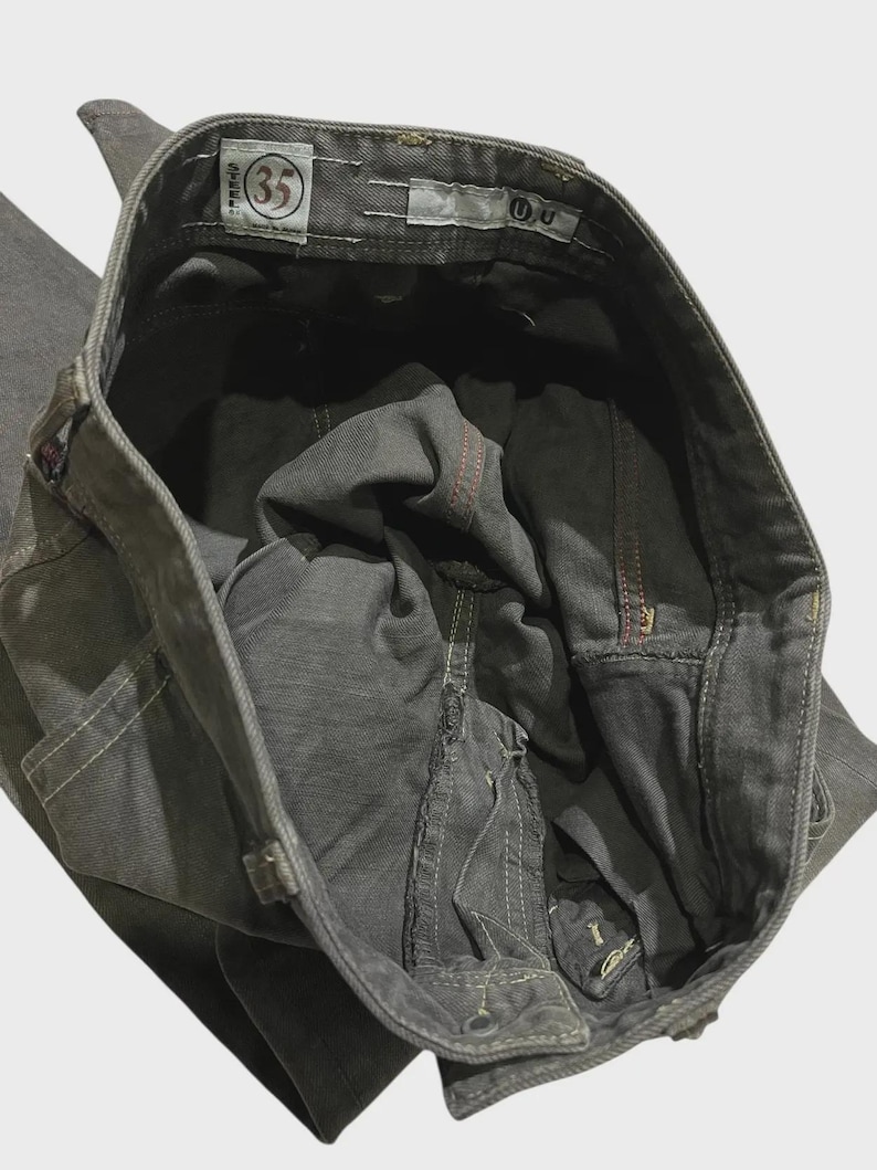 K&ouml;nnte beinhalten: Nahaufnahme einer grauen Jeans. Die Jeans hat einen Knopfverschluss und einen Rei&szlig;verschluss. Das Gr&ouml;&szlig;enetikett lautet "35". Die N&auml;hte sind in wei&szlig;em und rotem Faden sichtbar. Die Jeans ist gefaltet und zeigt die Innentaschen und den Bund.