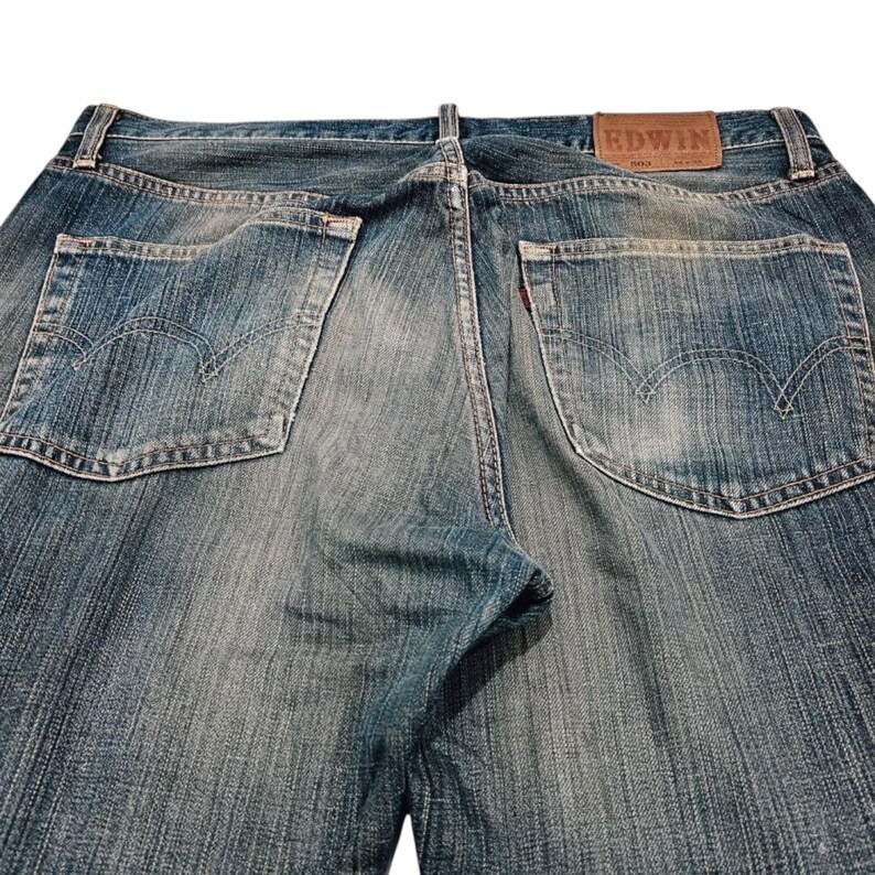 Puede incluir: Vaqueros de mezclilla azul descolorido con estilo cl&aacute;sico de cinco bolsillos. Incluye un parche de cuero marr&oacute;n con la marca "EDWIN" y una etiqueta roja en el bolsillo trasero. Los vaqueros tienen un aspecto desgastado.