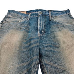 Puede incluir: Primer plano de unos vaqueros de mezclilla azules con un lavado descolorido. Los vaqueros tienen un dise&ntilde;o cl&aacute;sico de cinco bolsillos, cierre de bot&oacute;n y cremallera, y trabillas. La mezclilla tiene un aspecto desgastado.