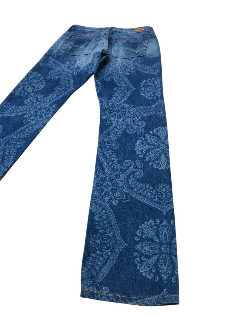 May include: Blue denim jeans with a light blue paisley pattern. The jeans have a classic five-pocket design and a straight leg. The back pockets and waistband are visible.