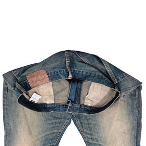Puede incluir: Vaqueros de mezclilla azul con un lavado descolorido. Los vaqueros est&aacute;n planos, mostrando los bolsillos interiores y un parche de cuero con texto en relieve. Una peque&ntilde;a etiqueta es visible cerca del bolsillo. Estilo cl&aacute;sico.