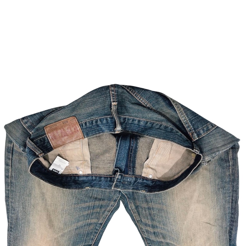 Puede incluir: Vaqueros de mezclilla azul con un lavado descolorido. Los vaqueros est&aacute;n planos, mostrando los bolsillos interiores y un parche de cuero con texto en relieve. Una peque&ntilde;a etiqueta es visible cerca del bolsillo. Estilo cl&aacute;sico.