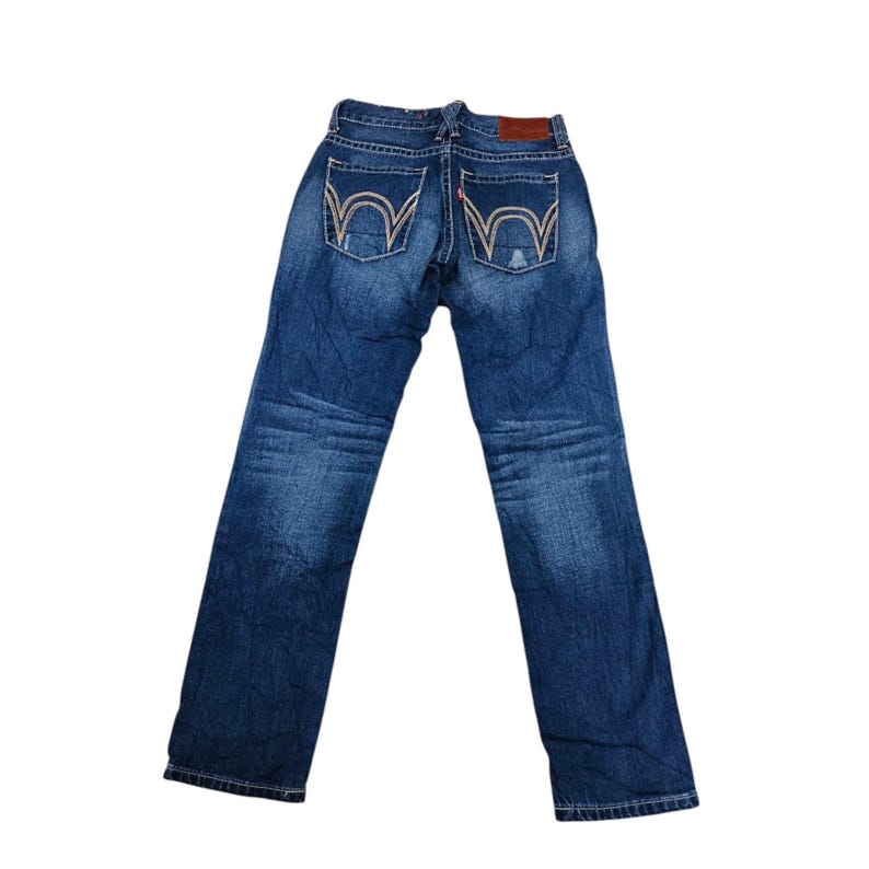 May include: Blue denim jeans with light stitching and a brown leather patch. The back pockets feature a distinctive design. The jeans are laid flat against a white background.