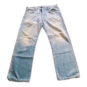 May include: Light blue denim jeans featuring a faded wash, a button fly closure, and a distressed worn-in texture with whiskering patterns across the upper thighs and a small frayed hole on the left knee area, finished with a straight leg cut.
