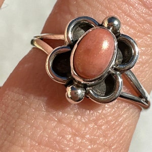May include: A silver ring with a pink oval gemstone at its centre, set within a floral design. The ring has a split shank and small silver ball accents. The gemstone is a light peach colour.