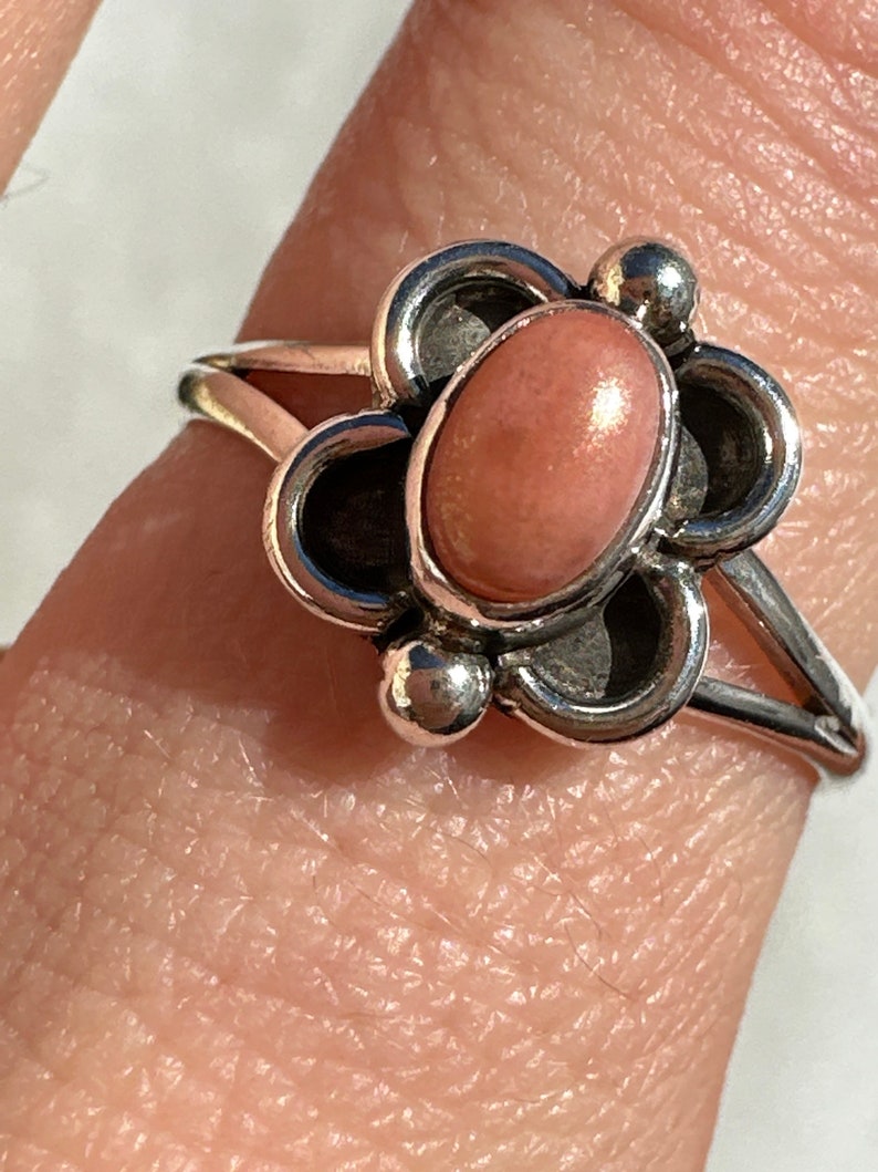 May include: A silver ring with a pink oval gemstone at its centre, set within a floral design. The ring has a split shank and small silver ball accents. The gemstone is a light peach colour.