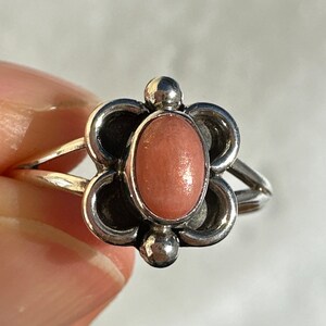 May include: A silver ring with a flower-shaped setting and a pink oval stone. The ring has a split band and small silver ball accents. The ring is a piece of jewellery.