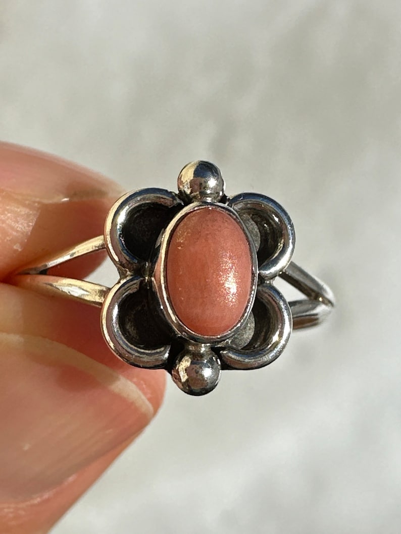 May include: A silver ring with a flower-shaped setting and a pink oval stone. The ring has a split band and small silver ball accents. The ring is a piece of jewellery.