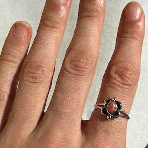 May include: A silver ring with a flower design, featuring a central oval peach-coloured stone. The flower has black petals and small silver accents. The ring is on a finger.