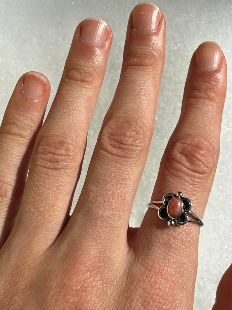 May include: A silver ring with a flower design, featuring a central oval peach-coloured stone. The flower has black petals and small silver accents. The ring is on a finger.