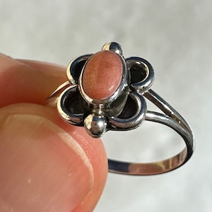 May include: A silver ring with a pink oval gemstone set in a floral design. The ring has a delicate band and small silver accents. The ring is held by a person's fingers.