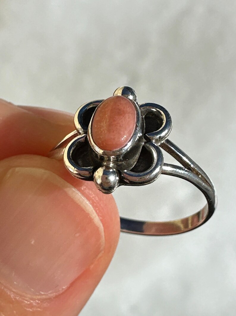 May include: A silver ring with a pink oval gemstone set in a floral design. The ring has a delicate band and small silver accents. The ring is held by a person's fingers.