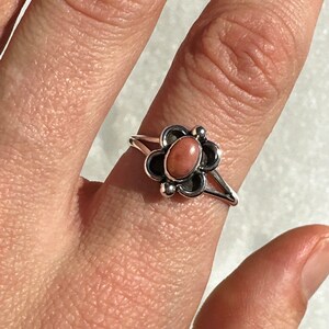 May include: A silver ring with a flower design and a pink gemstone centre. The ring has a detailed, openwork design with a split shank. The gemstone is a soft, muted pink colour, set in a dark metal flower setting.