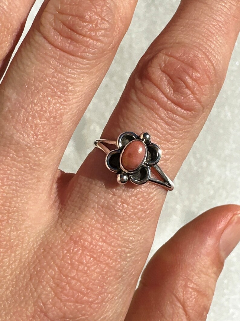 May include: A silver ring with a flower design and a pink gemstone centre. The ring has a detailed, openwork design with a split shank. The gemstone is a soft, muted pink colour, set in a dark metal flower setting.