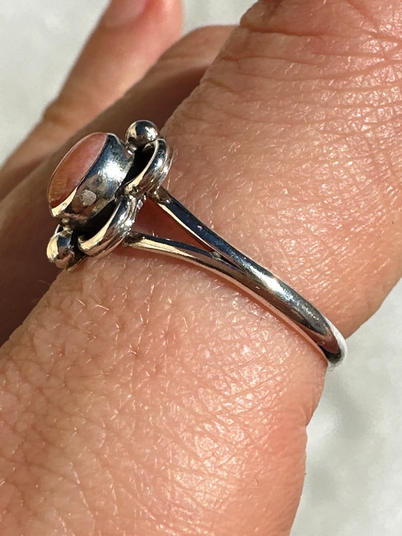 May include: A silver ring featuring a pink oval gemstone. The ring has a floral design around the stone and a smooth, polished band. The ring is shown on a finger.