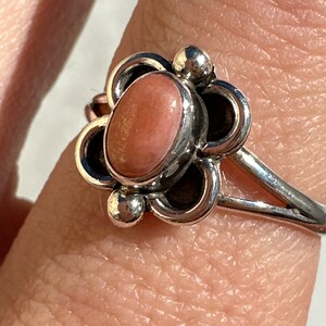 May include: A silver ring with a floral design, showcasing a central oval peach-coloured stone. The ring has a slender band, and the flower petals are detailed with small, round accents. The ring is displayed on a finger.