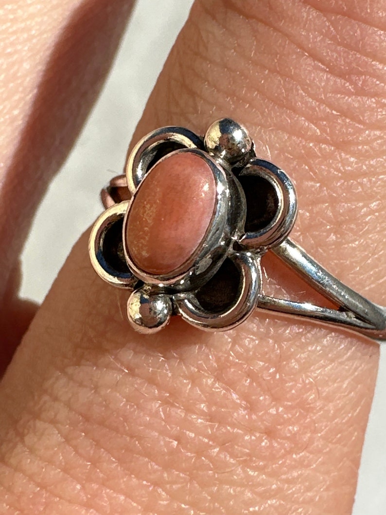 May include: A silver ring with a floral design, showcasing a central oval peach-coloured stone. The ring has a slender band, and the flower petals are detailed with small, round accents. The ring is displayed on a finger.