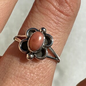 May include: A silver ring with a floral design, featuring a central oval pink stone. The ring has a slender band and decorative elements around the stone, offering an elegant and feminine aesthetic.
