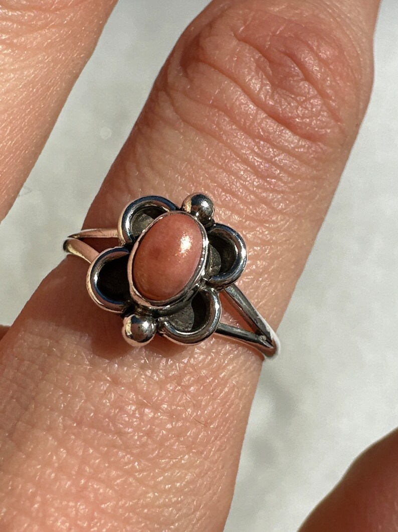 May include: A silver ring with a floral design, featuring a central oval pink stone. The ring has a slender band and decorative elements around the stone, offering an elegant and feminine aesthetic.