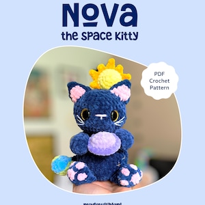 Könnte beinhalten: Ein dunkelblaues Häkel-Katzenspielzeug namens Nova the Space Kitty mit großen schwarzen Augen, rosa Innenohren und rosa Pfotenballen. Es hält eine hellviolette Kugel, hat ein gelbes Sonnenaccessoire hinter dem Kopf und einen bunten Schwanz.