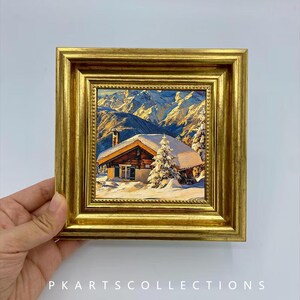 Pode incluir: Uma pequena pintura quadrada numa moldura dourada decorativa representando uma cabana de madeira coberta de neve, situada em frente a montanhas nevadas sob um céu azul claro. O texto PKARTSCOLLECTIONS está impresso em branco na parte inferior.