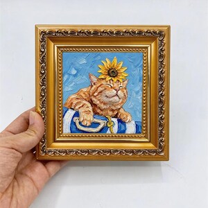 May include: An oil painting in a decorative gold frame featuring an orange tabby cat with a sunflower on its head. The cat is resting its paws on a blue and white striped object against a textured light blue background. The painting is signed.