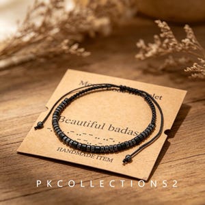 Peut inclure: Un bracelet en cordon noir avec de petites perles métalliques disposées en code Morse. Le bracelet est présenté sur une carte beige portant le texte Beautiful badass et HANDMADE ITEM, ainsi qu'une séquence en code Morse.
