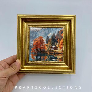 Pode incluir: Uma pequena pintura de paisagem quadrada numa moldura dourada espessa e ornamentada. A obra retrata uma cena de lago de montanha com árvores de outono laranja vibrantes, uma pequena cabana e picos rochosos refletidos na água. O texto PKARTSCOLLECTIONS é visível.