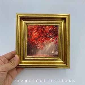 May include: A square framed print featuring a vibrant autumn forest scene with bright red leaves and sunbeams filtering through the trees. The artwork is encased in a thick, ornate gold-colored frame with a beaded inner edge and a smooth outer border.