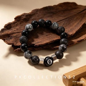 Könnte beinhalten: Ein Perlenarmband mit einer Mischung aus schwarzen und grauen kugelförmigen Perlen, darunter glatte, poröse und gemusterte Varianten. Das Armband enthält zwei metallische, silberfarbene quadratische Zwischenstücke und eine schwarze Perle mit der weißen 8.