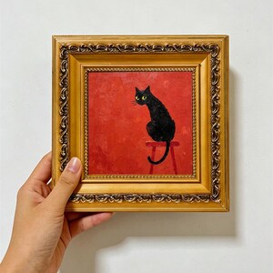 May include: A square oil painting in a decorative gold frame featuring a black cat with yellow eyes sitting on a red stool against a solid red background. The text PKARTSCOLLECTIONS is printed in white at the bottom of the image.