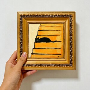 May include: A square oil painting in an ornate gold frame depicting a black cat resting on a set of yellow stairs. The painting features visible brushstrokes and a warm color palette. The text PKARTSCOLLECTIONS is printed at the bottom of the image.