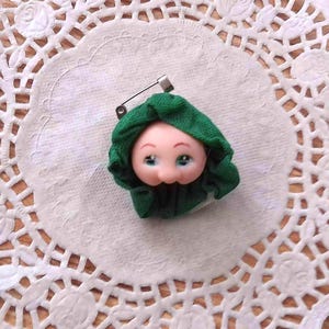 May include: A brooch featuring a small, pale-skinned doll head with painted facial features, framed by a gathered dark green fabric hood. The doll head is attached to a metal pin mechanism on the back, allowing it to be fastened to clothing.