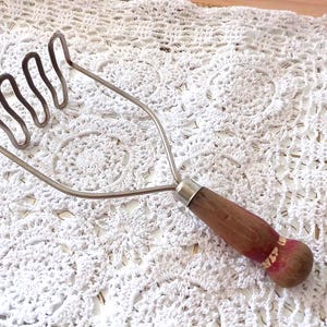 May include: A vintage potato masher with a wooden handle and a metal head. The handle is brown with a red and white accent near the end. The metal head has four looped wires.