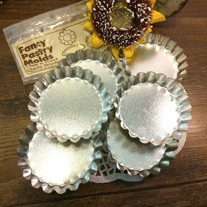 May include: A collection of silver-colored, fluted tart molds, with a package of "Fancy Pastry Molds" in the background. A decorative sunflower is also present. The molds are made in France and designed for quiche and brioche.