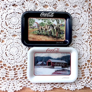 May include: Two vintage Coca-Cola trays. One is black with a rural scene, the other is white with a winter landscape. Both trays have the Coca-Cola logo in red and white. The trays are rectangular with rounded corners.